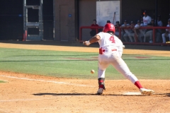 Baseball player wearing number 4 bunting the ball during a game, with the ball just leaving the bat and teammates watching from the dugout in the background.