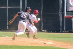 Palomar baseball player in white and red turns to run towards second base as the throw to first missed and the Saddleback player in black runs to retrieve the overthrown ball.