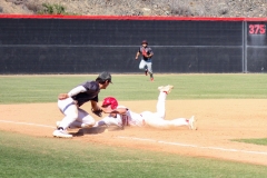 Baseball player in a red helmet dives headfirst toward third base as an opposing fielder reaches down to apply the tag, with another player running in from the outfield.