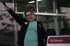 Anel Gonzalez is wearing a black sweater and a green CCE shirt with a CCE pin, holding a microphone up for the audience to repeat her version of "Baby It's Cold Outside, replacing the lyrics with  "Baby it's COLA outside.
