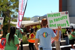 a man in a blue PFF shirt holds a "invest in faculty and staff!" sign while looking on to a woman in an orange vest making a speech