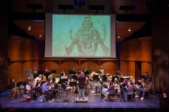 A group of musicians is sitting on stage performing while a director in a black t-shirt conducts them. A screen above them is projecting drawings of different cultures from around the world.