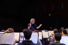 A woman in a black t-shirt and glasses is holding a conductor’s baton in one hand and has her other arm raised in front of a group of musicians on stage.