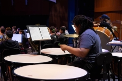 A man in a blue t-shirt is reading sheet music off of the stand in front of him while playing the timpani on stage.