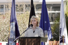 A blond woman in a beige jacket stands behind a podium, giving a welcome speech at the Veterans Day Ceremony.