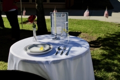 A table with a white cloth is set up in the Veterans Memorial Lawn. On the table there is a red rose, a plate with a lemon, a white candle, upside down wine glass, and utensils.