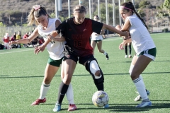 Taylor Devaney (#16) in black, has the soccerball and is surrounded by two Grossmont players in white, who are trying to steal the ball from Devaney.