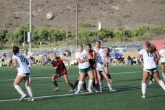 Sydney Kemp (#2) and Sophia Federick (#12), both in black, are surrounded by players in white uniforms from Grossmont College as they try to retrieve the soccer ball after a corner kick.