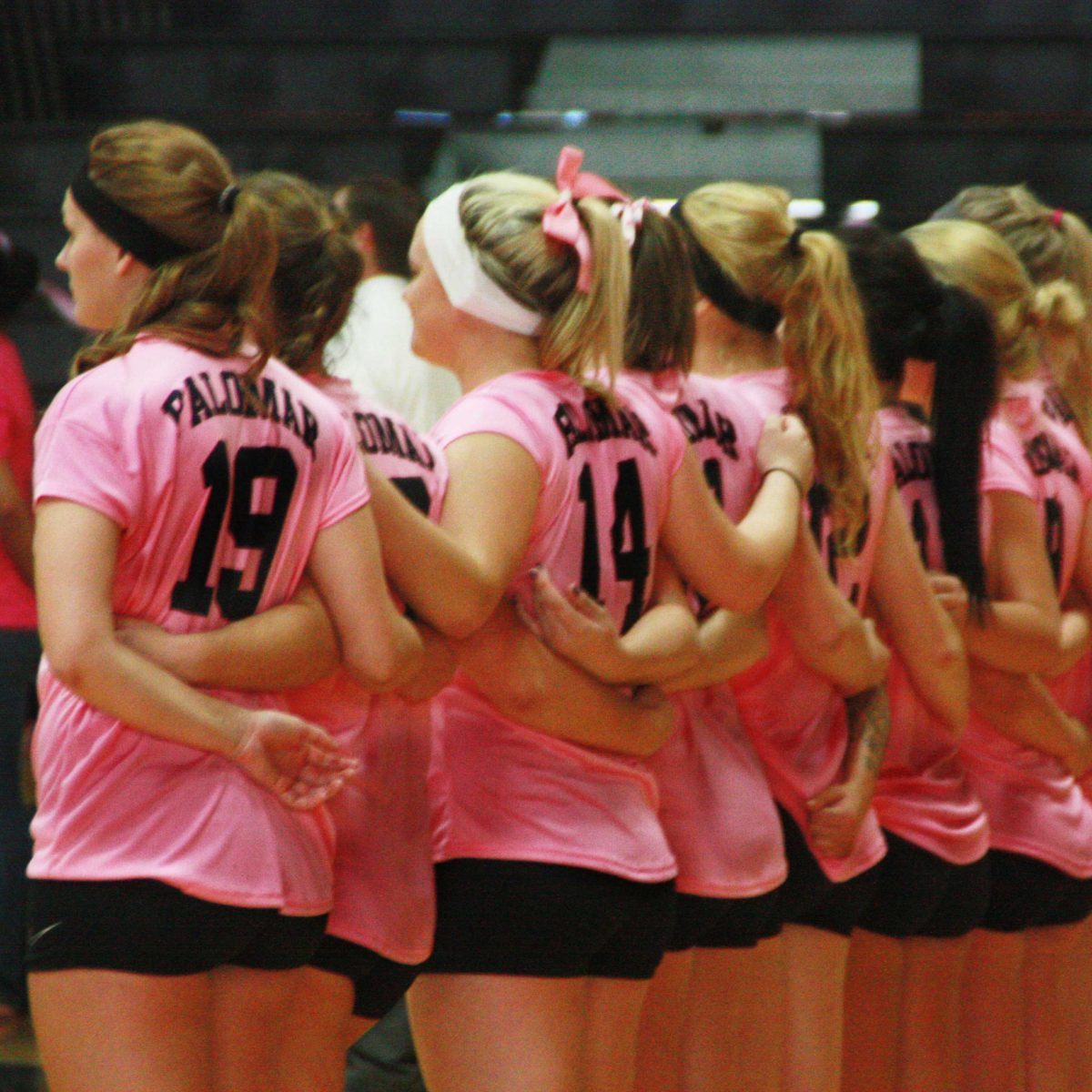 A team of female Palomar volleyball players hold each other around the waist in a line before their match. Almost all wear pink jerseys and black shorts.