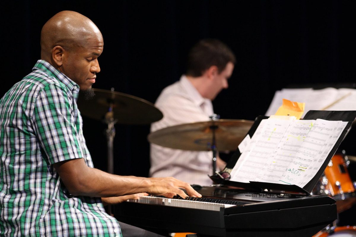 A man plays a piano with a (blurred) man playing the drums in the background.