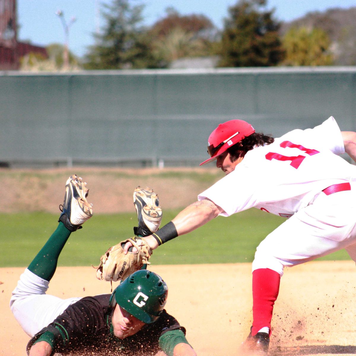 A Palomar baseball player leans foward and tags an opposing player with his mitted left hand. The other player slides on his stomach, wearing a green helmet, black jersey, white pants, and long green socks.