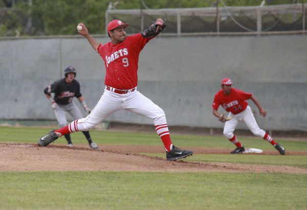 A male Palomar baseball player prepares to throw a baseball with his right hand, his right leg extended behind him. Another team player on the right prepares to bolt to another base as an opposing player on the left looks on.