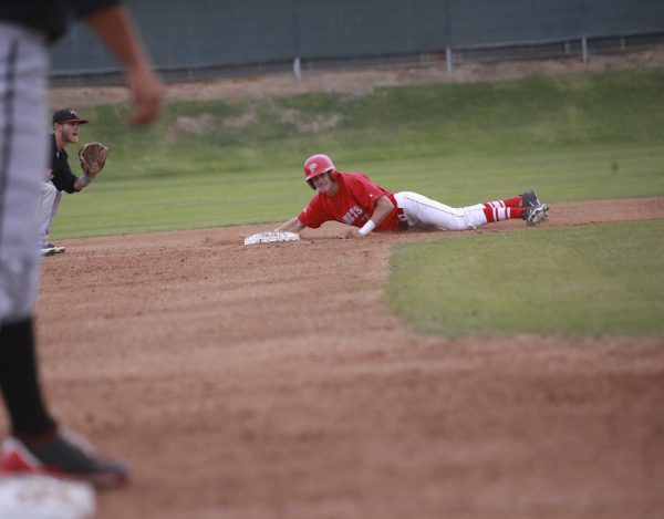 A Palomar baseball player slides on the ground on his stomach toward a base. A right leg of an opposing player is on the left. Another opposing player in the background near the base readies his left-mitted hand to catch a baseball.
