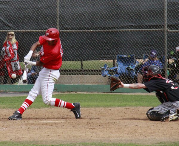 A male Palomar baseball player swings a bat toward an incoming baseball. A catcher squats behind him with his left-mitted hand extended to catch the ball. A few people sit or stand behind a chain-linked fence watching the game.