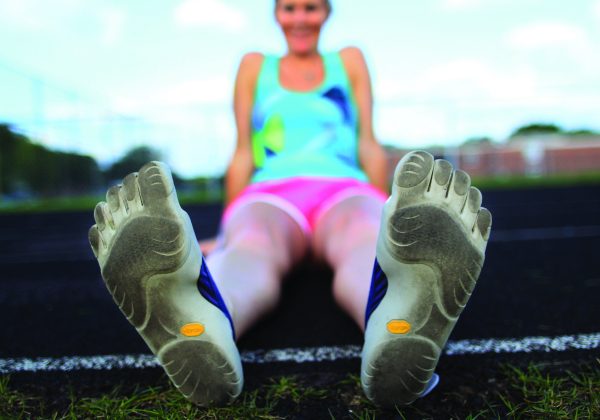 A woman sits on the ground with the bottom of her feet, which is wearing Vibrams, facing the viewer.
