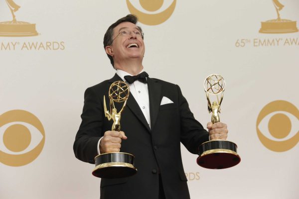 Comedian Stephen Colbert stands and laughs, holding an Emmy Award trophy in each hand with his body tilted slightly back.