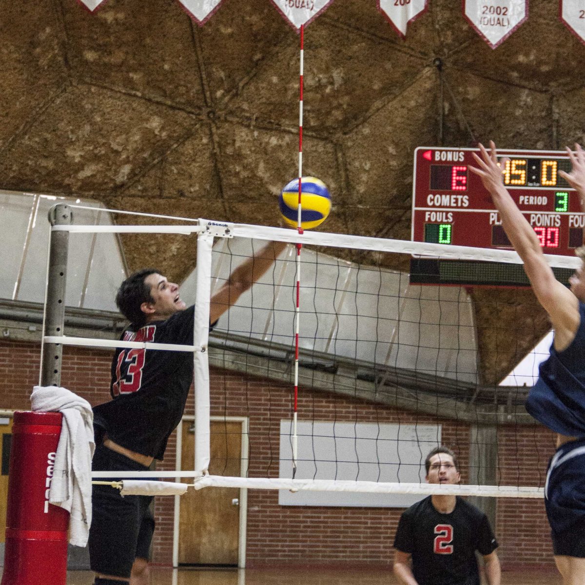 A male Palomar volleyball player jumps and hits the ball over the net as an opponent on the right tries to block it.