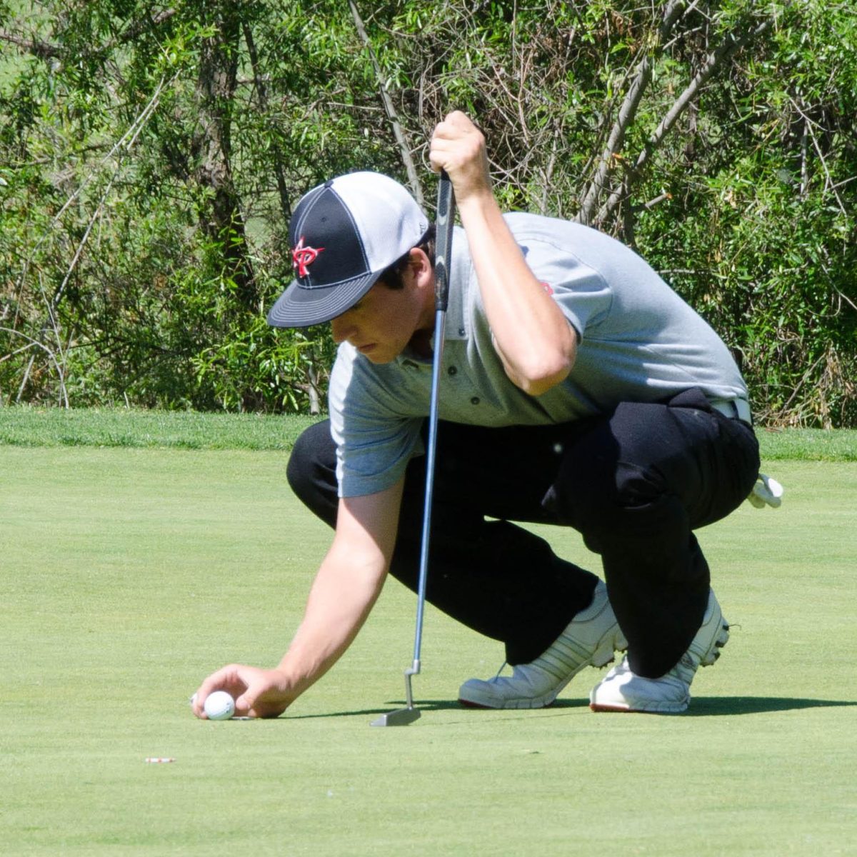 A male Palomar golfer crouches and puts a golf ball on the turf. He holds a golf club in his left hand propped up on the ground.