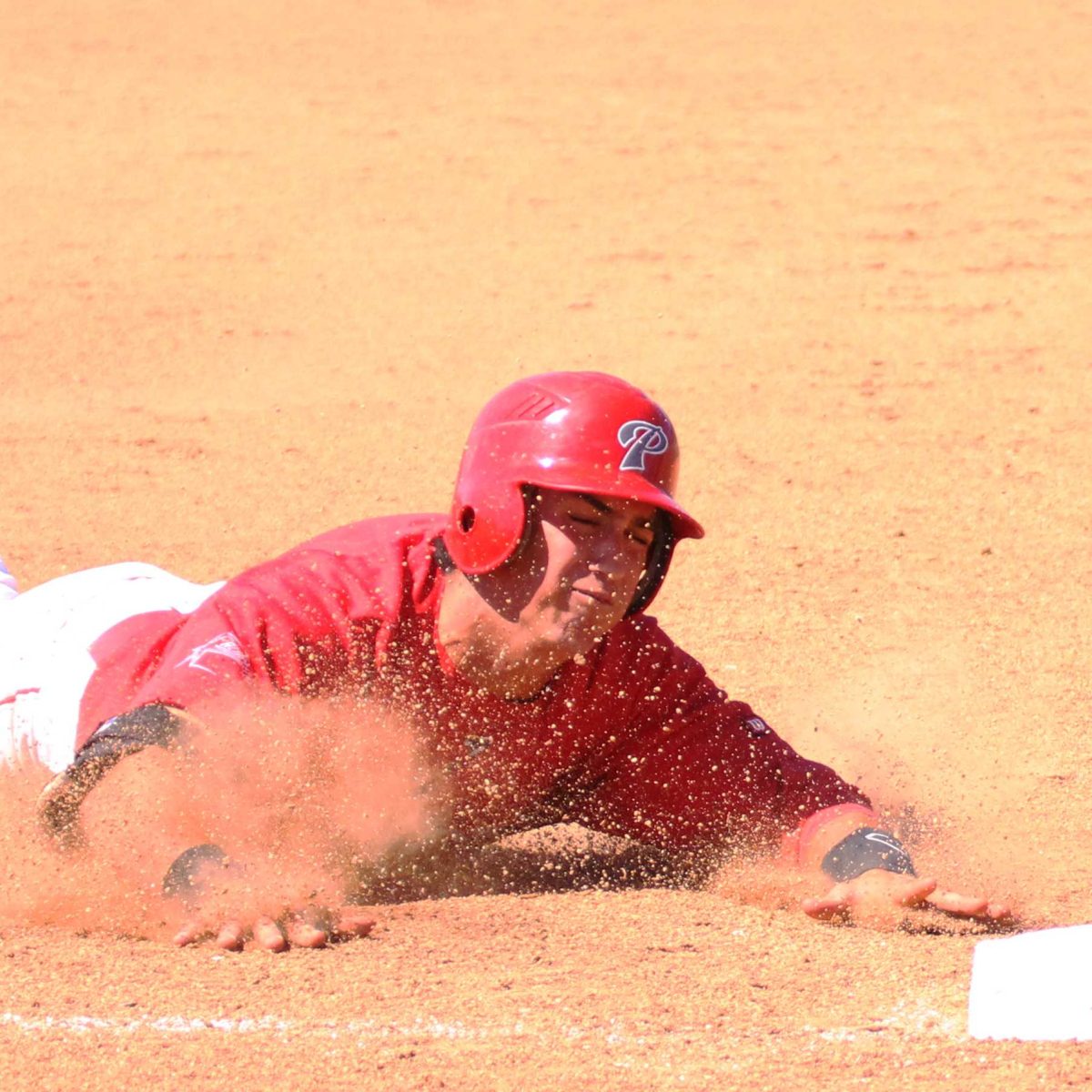 A male Palomar baseball slides to base on his stomach, dirt flying in front of him.