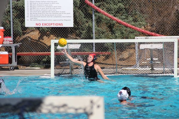 A female Palomar water polo goalie tries to block a ball from entering a goal with her right hand.