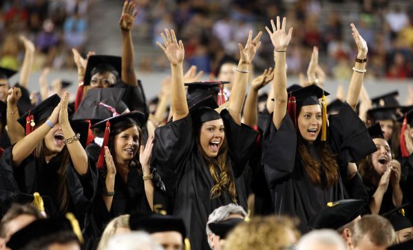 Four female students cheer during a graduation ceremony. Two on the right raise their hands above their heads. The two on the left clap their hands. A crowd of people are around them.