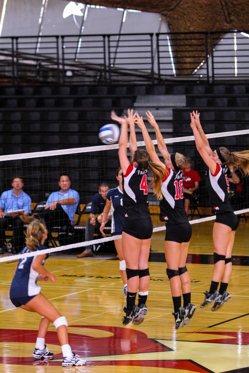 Three female Palomar volleyball players try to block the ball after an opponent hits it over the net.