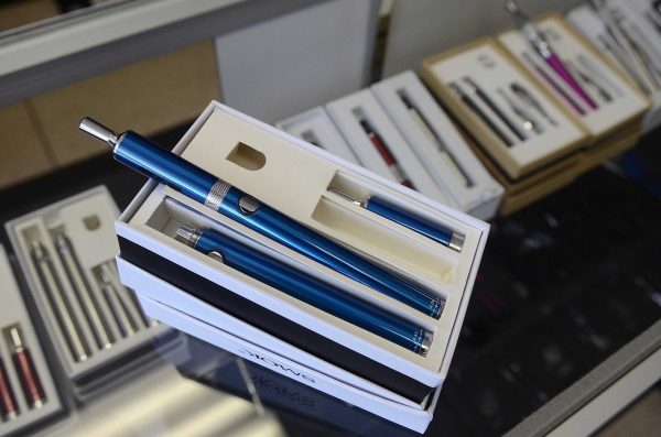 A blue e-cigarette inside a small white box, sitting on top of a glass case with more e-cigarettes in small white boxes.