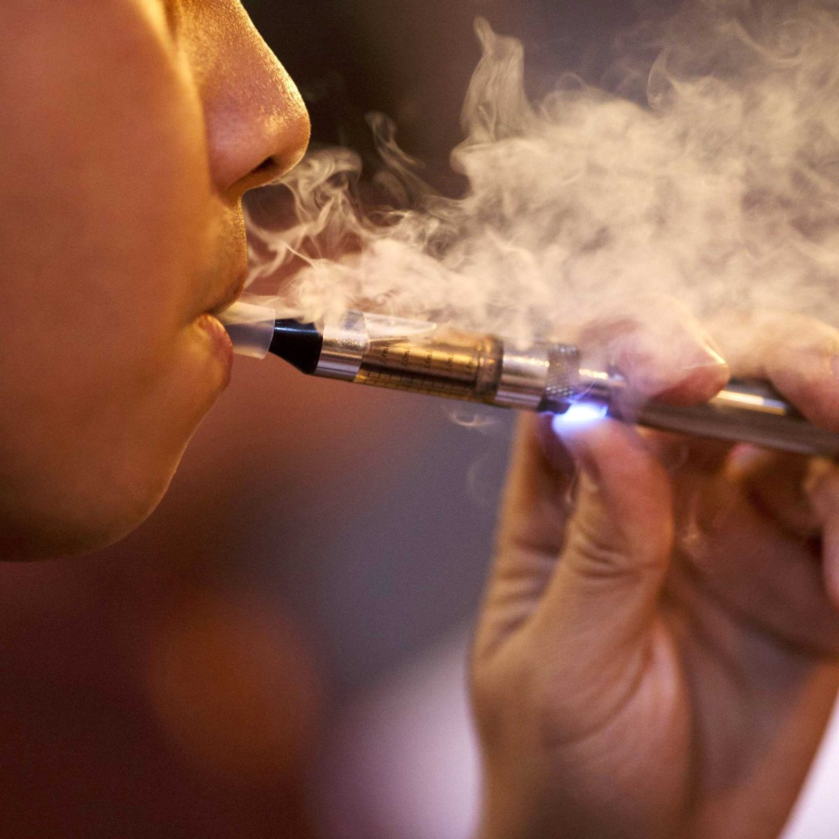 A man is smoking an e-cigarette, holding it with his left hand. Smoke puffs out from his mouth.