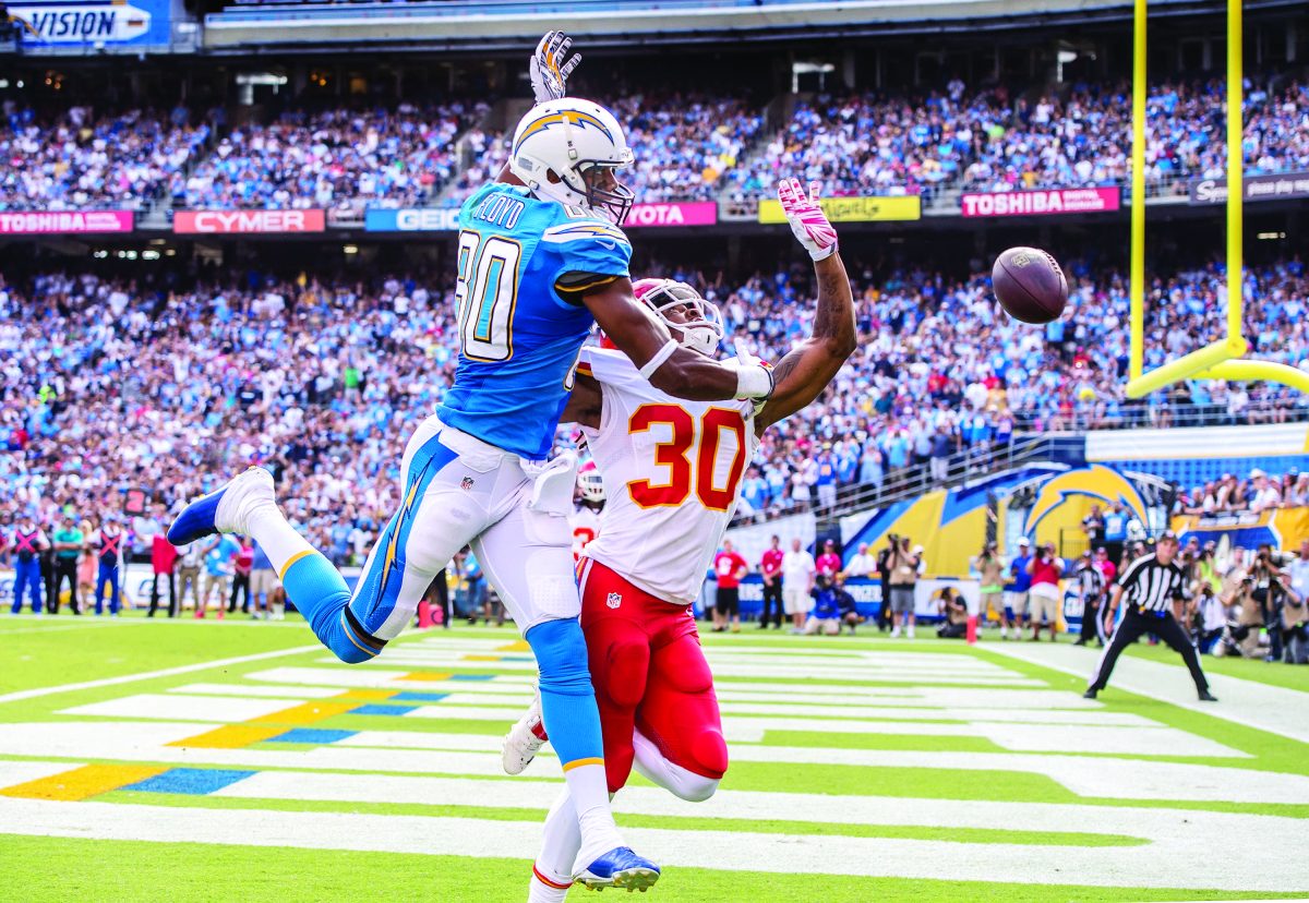 A football player in a bright blue jersey and white pants tackles another player in a white jersey and red pants. A football flies to the right. A referee watches in the background on the right, and a huge crowd fill the stadium stands in the background.