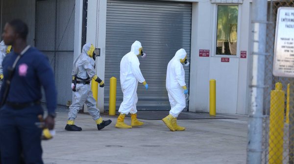 Three people wearing a Hazmat suit walk in a queue toward a building entrance. A man with a blue long-sleeve shirt and pants stand to the left in the foreground (blurry).
