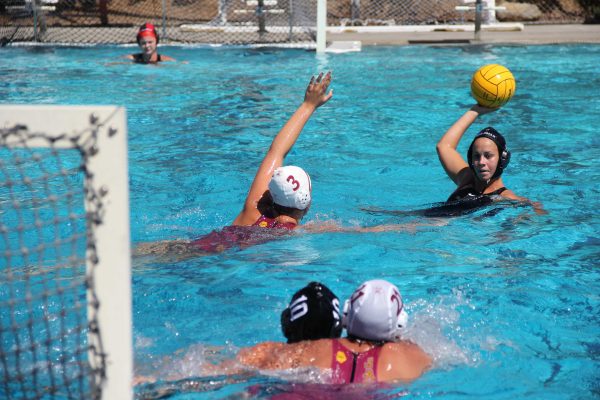 A female Palomar water polo player throws a ball toward the goal with her right hand. An opponent tries to block her.
