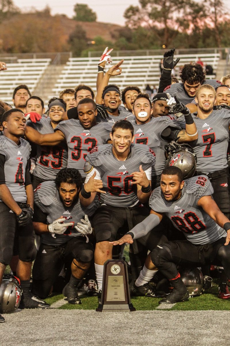 A team of Palomar football players gather and pose for a picture after they won a game. A trophy sits on the ground in front of them.