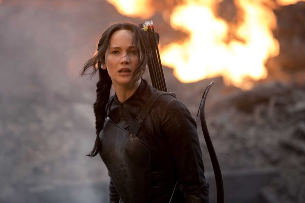 Actress Jennifer Lawrence as Katniss Everdeen in the film "The Hunger Games: Mockingjay Part 1." She hold a bow and carries a quiver of arrows. A fire burns in the background.