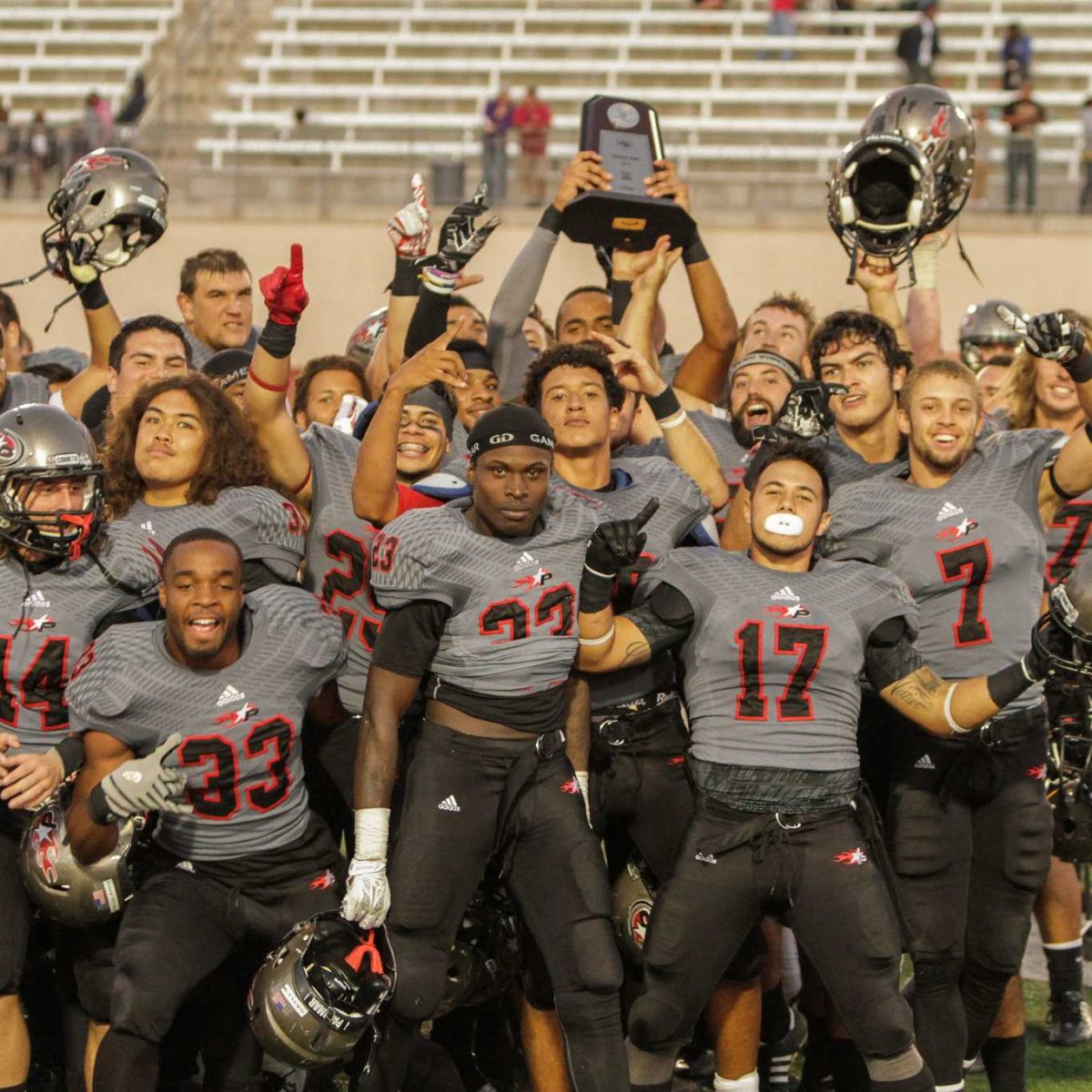 A team of Palomar football players gather and pose for a picture after they won a game. Several in the background hold up their helmets and a trophy.