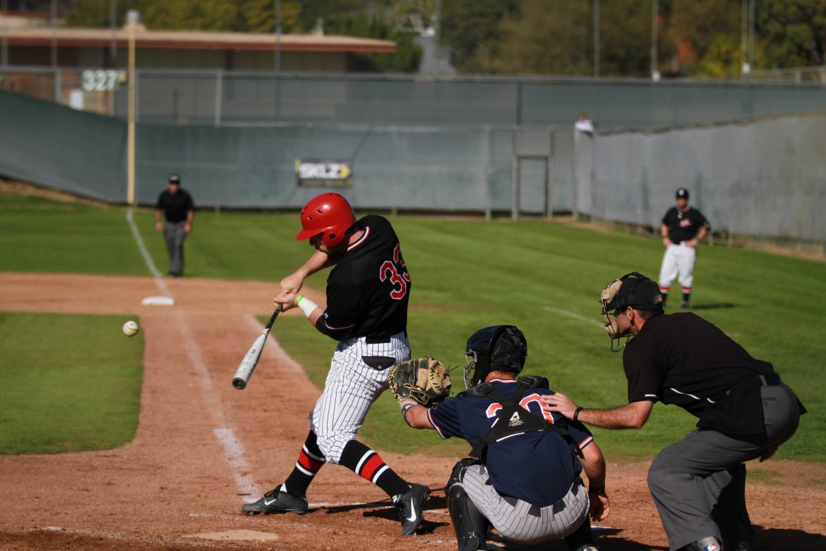 A male Palomar baseball player swings a bat toward an incoming baseball. A catcher squats behind him with his left-mitted hand extended to catch the ball. An umpire stands behind the catching, watching the ball. In the background, an umpire stands on the left and another team player stands on the right.