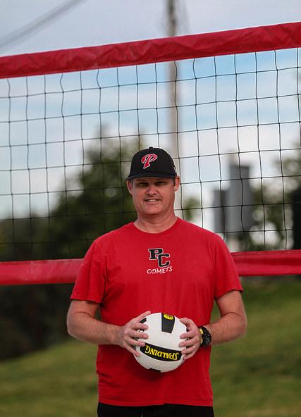 Women's sand volleyball head coach Karl Seiler holds a volleyball with both hands, wearing a black cap and red T-shirt. A volleyball net is behind him.