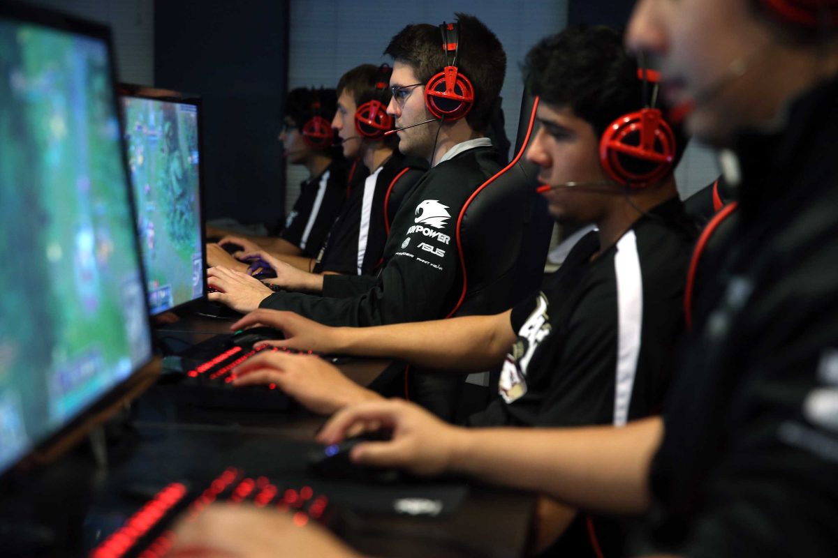 A group of young men sit in front of computer monitors and keyboards, wearing headphones.