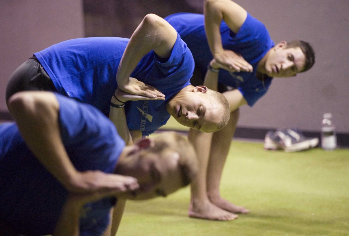 Three men perform a yoga pose with their feet and palms together, their body bent forward, and turned to their right.