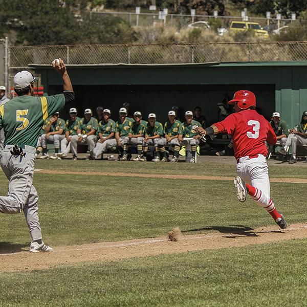 A male Palomar baseball player runs to the right, looking back at the opposing player who is about to throw a baseball toward him. Many opposing players sit side by side on a bench in the background near a dugout.