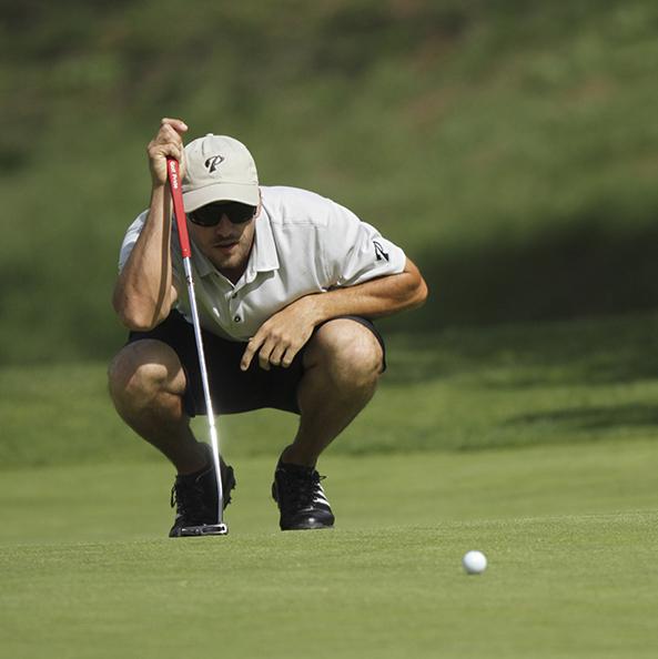 Palomar golfer Nick Bellstrom crouches and looks at a golf ball a few feet in front of him. He holds a golf club propped up on the ground in his right hand.