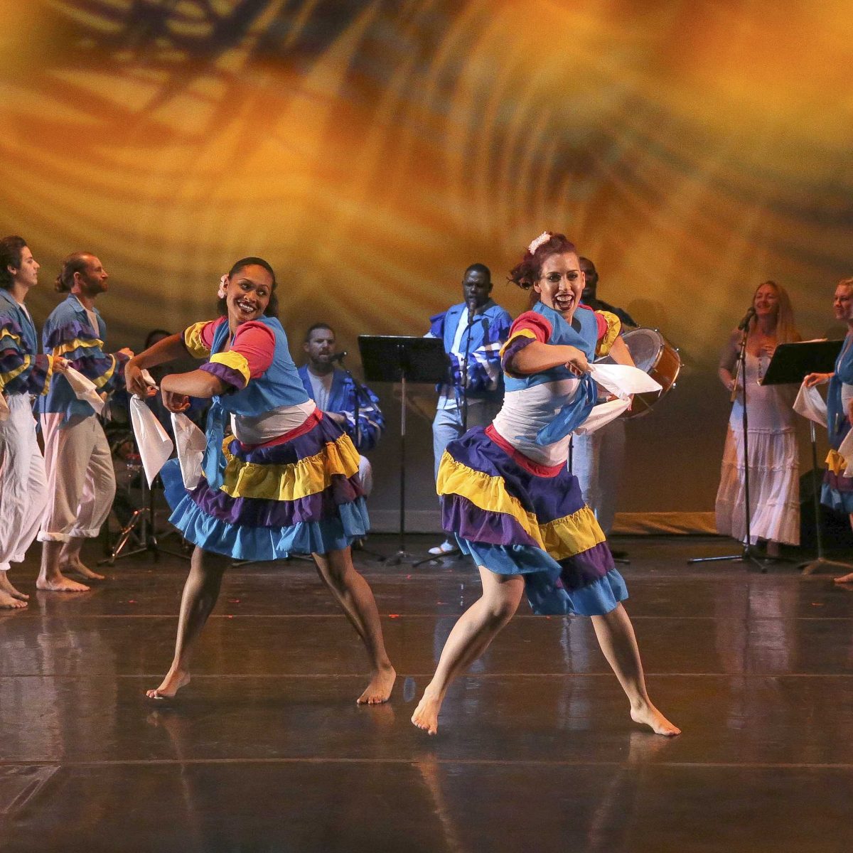 Two female dancers wearing Latin-style dresses in blue, purple, yellow, white, and red dance on a stage with several other dancers and musicians in the background.