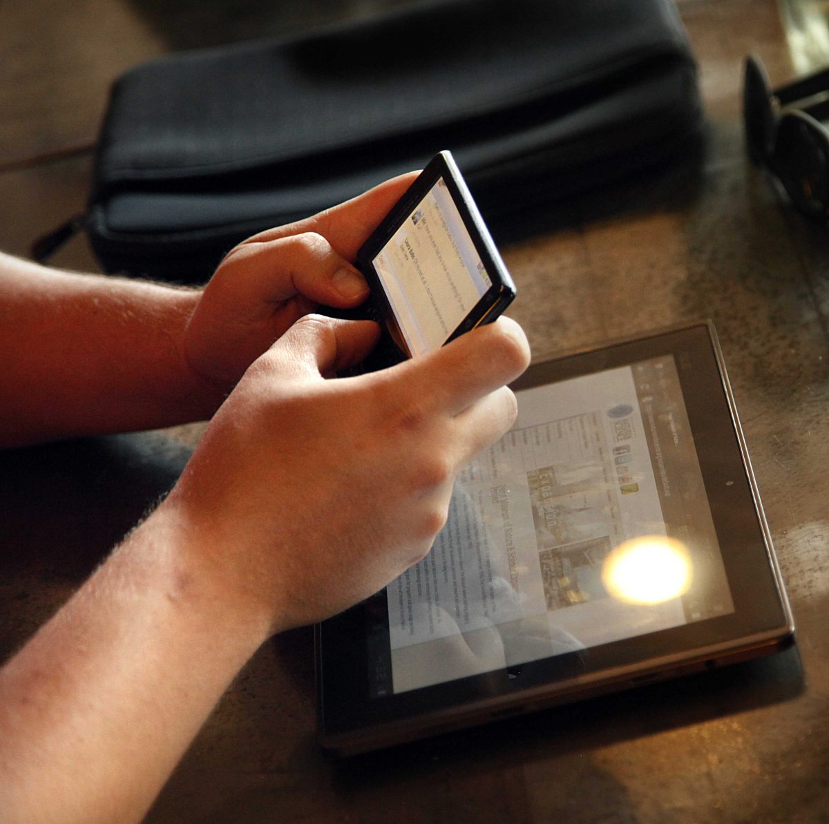 Two hands hold an Android phone with a Tablet on a table.