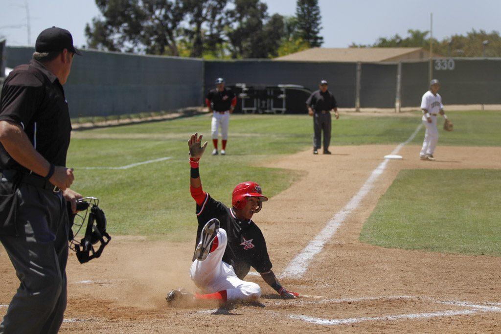 A Palomar baseball players to a base on his bottom, yelling with his right hand up in the air. An umpire stands and looks at him on the left. Another Palomar player, an umpire, and an opposing player stand in the background and watch him.