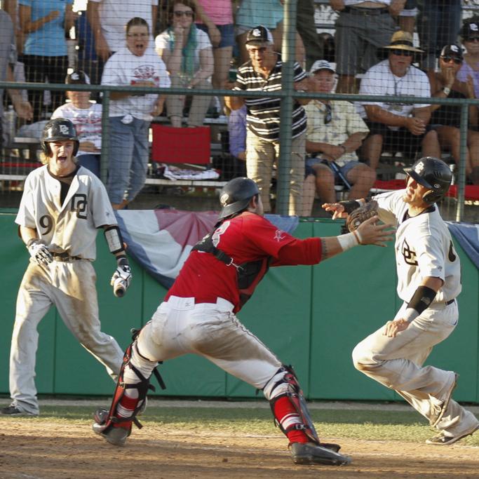 A male Palomar baseball player tags an opposing player on the right as another player walks and yells on the left. Fans watch with excitement in the background behind a net.