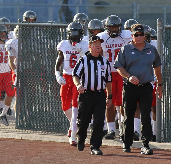 Palomar head coach Joe Early walks with a referee and the team follows them through a metal gate.