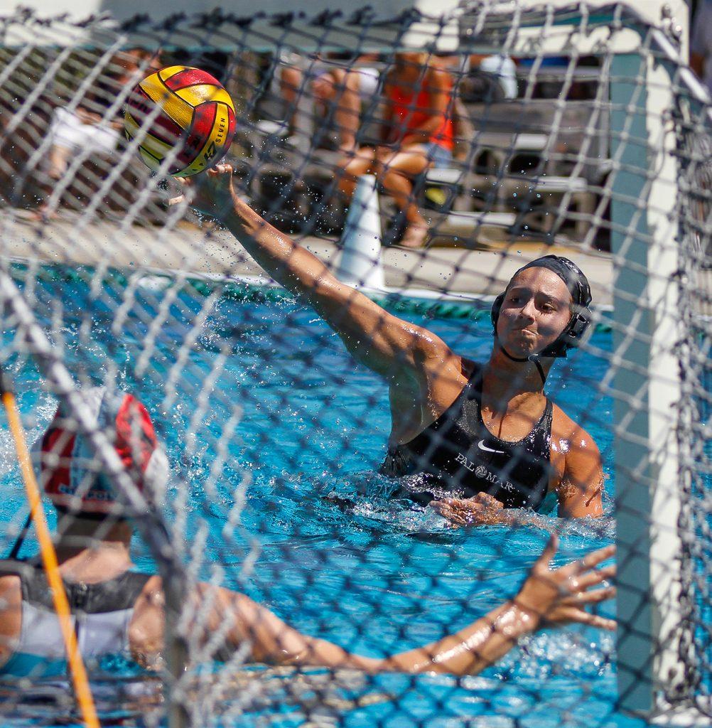 A female Palomar water polo player throws a ball toward the goal with her right hand. An opponent is at the goal.