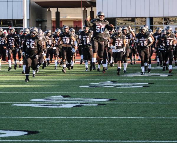 A team of Palomar football players runs across a field as one of them in the middle leaps in the air, cheering.