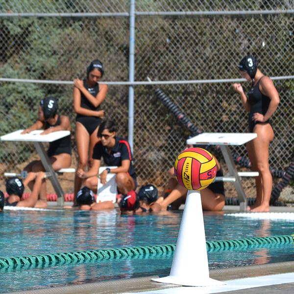 A water polo ball sits atop of a white plastic cone at the edge of a pool. Water polo coach Jem McAdams crouches near the pool and speaks to her team.