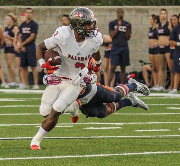 A Palomar football player runs with a football tucked in his right arm as an opposing player tries to tackle him from behind and falls. Several opposing players and cheerleaders stand in the sidelines in the background.