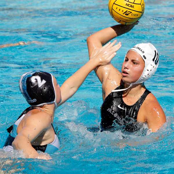 A female Palomar water polo player tries to throw the ball in her right hand as an opponent tries to block her from the left.